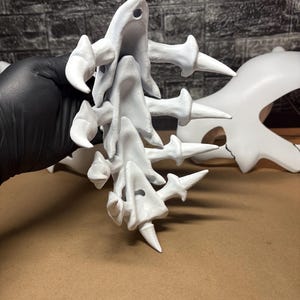 May include: A collection of white, abstract 3D-printed sculptures. The forms have a bone-like appearance with sharp, pointed projections and rounded, curved elements. The pieces are held against a neutral background.