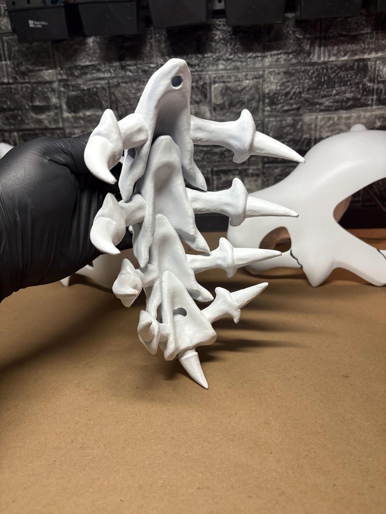 May include: A collection of white, abstract 3D-printed sculptures. The forms have a bone-like appearance with sharp, pointed projections and rounded, curved elements. The pieces are held against a neutral background.