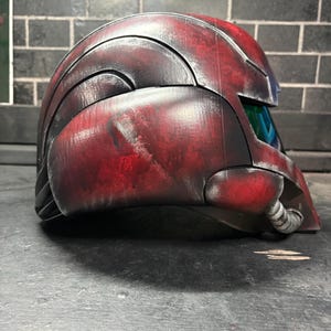 May include: A side profile of a full-face helmet featuring a weathered red and black finish. The helmet has a curved, segmented shell design with a dark, reflective visor and a small, ribbed circular attachment near the jawline area.