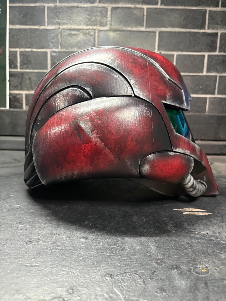 May include: A side profile of a full-face helmet featuring a weathered red and black finish. The helmet has a curved, segmented shell design with a dark, reflective visor and a small, ribbed circular attachment near the jawline area.