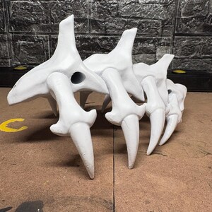May include: A white sculpture of a skeletal form. The piece has multiple bone-like components with sharp, pointed projections. The sculpture is set against a textured, dark grey background, with a brown surface in the foreground.