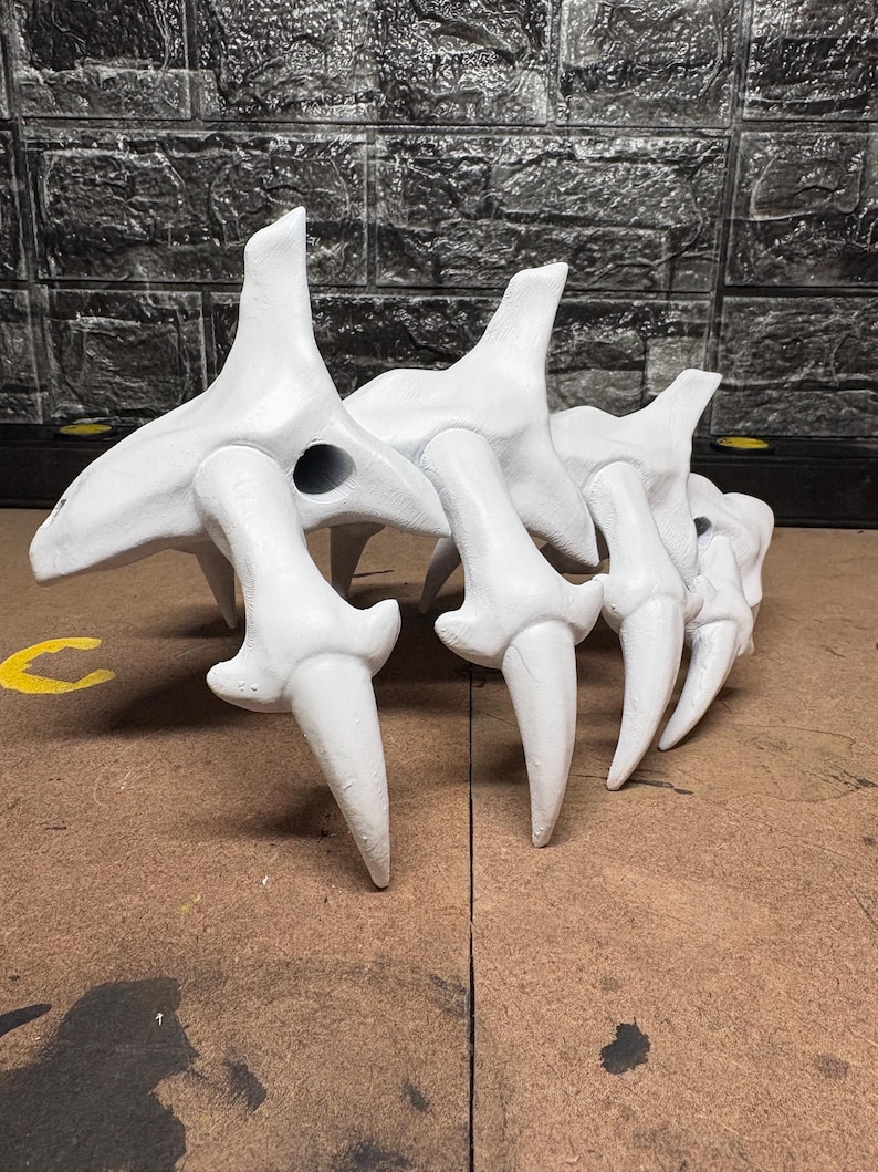 May include: A white sculpture of a skeletal form. The piece has multiple bone-like components with sharp, pointed projections. The sculpture is set against a textured, dark grey background, with a brown surface in the foreground.