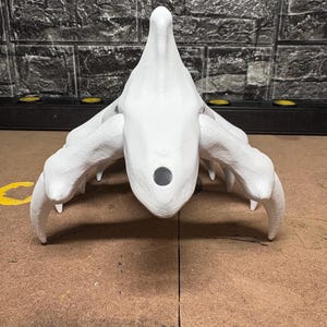May include: A white, abstract sculpture with a central, rounded form and multiple curved, claw-like appendages extending outwards. The sculpture has a textured surface and a dark circular opening. The background features a textured, dark grey brick wall.