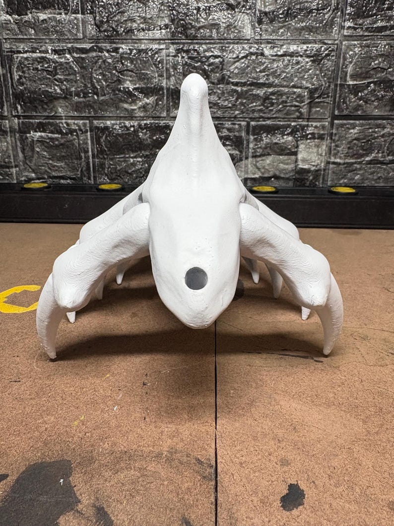May include: A white, abstract sculpture with a central, rounded form and multiple curved, claw-like appendages extending outwards. The sculpture has a textured surface and a dark circular opening. The background features a textured, dark grey brick wall.