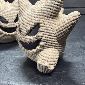 Pode incluir: Duas figuras de croch&ecirc; em forma de criatura redonda e espinhosa com orelhas pontudas. As figuras s&atilde;o feitas de um cord&atilde;o grosso bege claro com inser&ccedil;&otilde;es de tecido preto formando os olhos e uma boca larga, irregular e sorridente.