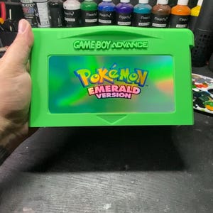 May include: A green Game Boy Advance cartridge with the text "GAME BOY ADVANCE" at the top. The cartridge features the "Pokémon Emerald Version" logo in the center. Paint bottles are visible in the background.