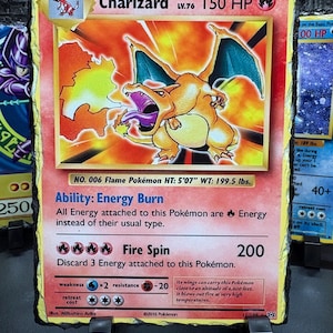 May include: A Charizard Pokémon card, stage 2, with a fiery orange and yellow design. The card features a detailed illustration of Charizard, a flame Pokémon, with the text "Fire Spin" and "200". The card is mounted on a black stand.