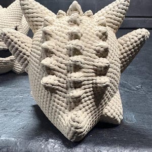 Pode incluir: Uma figura de croch&ecirc; na cor creme com corpo arredondado, m&uacute;ltiplos espinhos ao longo do topo e das laterais, e uma superf&iacute;cie tecida texturizada. O objeto apresenta uma silhueta sim&eacute;trica e pontiaguda com uma constru&ccedil;&atilde;o densa de fios.