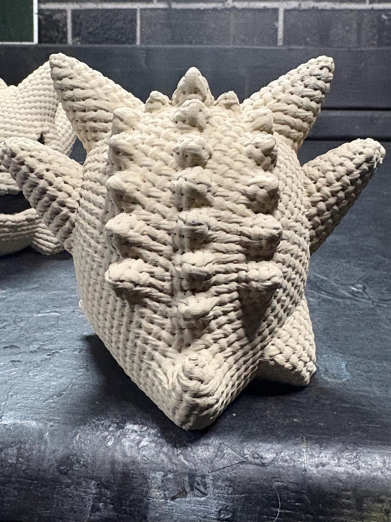 Pode incluir: Uma figura de croch&ecirc; na cor creme com corpo arredondado, m&uacute;ltiplos espinhos ao longo do topo e das laterais, e uma superf&iacute;cie tecida texturizada. O objeto apresenta uma silhueta sim&eacute;trica e pontiaguda com uma constru&ccedil;&atilde;o densa de fios.