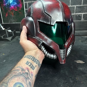 May include: A red and silver helmet featuring a dark green visor and metallic finish. The helmet has a segmented design with silver tubing details along the lower jaw area and a smooth, curved exterior shell with weathered, distressed textures.