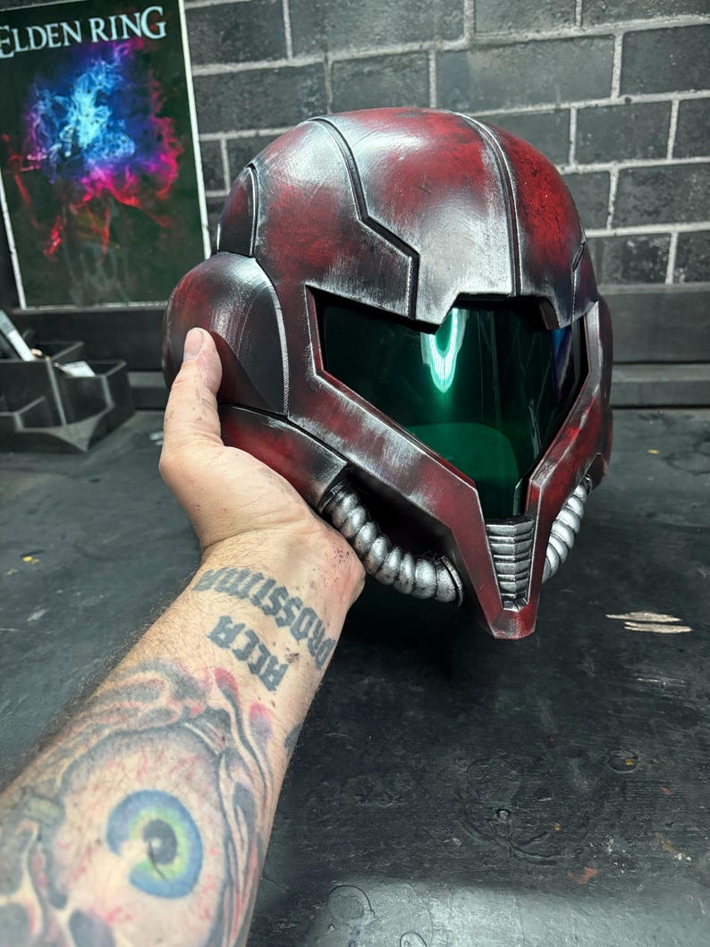 May include: A red and silver helmet featuring a dark green visor and metallic finish. The helmet has a segmented design with silver tubing details along the lower jaw area and a smooth, curved exterior shell with weathered, distressed textures.