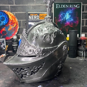May include: A silver helmet with intricate details and a blue and silver crest. The helmet is a replica of a helmet from the video game Elden Ring.