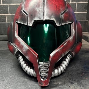 May include: A full-face helmet featuring a weathered red finish with silver metallic accents and a dark green reflective visor. The design includes two silver corrugated tubes extending from the chin area and angular paneling across the exterior.
