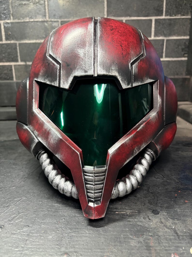 May include: A full-face helmet featuring a weathered red finish with silver metallic accents and a dark green reflective visor. The design includes two silver corrugated tubes extending from the chin area and angular paneling across the exterior.
