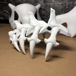 May include: White, stylized bone structures, possibly for a costume or display. The image shows a spine-like structure with pointed ends, and a skull-like mask in the background. The objects are on a brown surface.