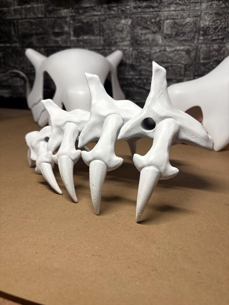 May include: White, stylized bone structures, possibly for a costume or display. The image shows a spine-like structure with pointed ends, and a skull-like mask in the background. The objects are on a brown surface.