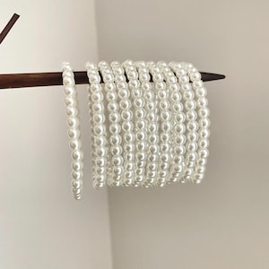 Puede incluir: Una colección de once pulseras de perlas blancas ensartadas en cordón elástico, dispuestas horizontalmente sobre una aguja de tejer de madera de color marrón oscuro. Cada pulsera consta de cuentas blancas redondas, uniformes y brillantes.