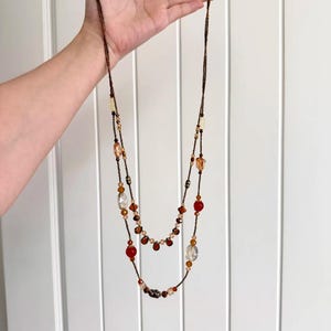Puede incluir: Un collar largo de varias hebras con cadenas de cuentas de color marrón oscuro, adornado con una variedad de cuentas translúcidas y opacas en tonos ámbar, transparentes y rojo intenso. El diseño incluye cuentas en forma de lágrima y redondas.