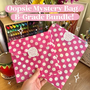 May include: Two pink polka dot paper bags labeled "Oopsie Mystery Bag/ B-Grade Bundle!" are held in front of a desk. The bags have white polka dots and a circular sticker that says "Oopsie Mystery Bag!". A bouquet of flowers is in the background.