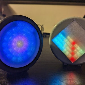 Puede incluir: Dos luminarias redondas, negras y blancas, con una matriz circular de LED. La primera luminaria tiene una luz azul y morada, mientras que la segunda luminaria tiene una luz azul, verde y roja.