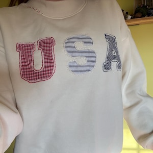 May include: A cream-colored crewneck sweatshirt featuring the letters USA appliqued on the front. The letter U is red and white gingham, the letter S is blue and white striped, and the letter A is blue and white gingham, all with visible stitching.