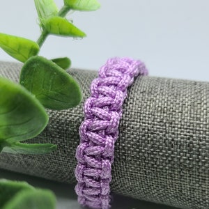 May include: A purple braided bracelet made of a soft, woven material. The bracelet is lying on a gray fabric surface.
