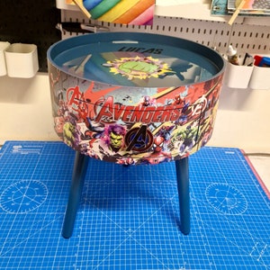 May include: A blue and red side table with a drawer featuring a graphic of the Avengers characters. The table has a round top and three legs. The top of the table is personalized with the name "Lucas".