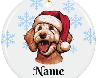 May include: A round, white Christmas ornament featuring a smiling dog wearing a red Santa hat. Light blue snowflakes are scattered around the dog's head. The word "Name" and the year "2024" are printed in black script below the dog's face. A gold string is attached for hanging.