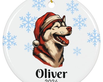 May include: A round, white ceramic ornament with a smiling husky wearing a red Santa hat. The ornament has the name "Oliver" and the year "2024" printed on it, with light blue snowflakes scattered around the edges. A gold string is attached for hanging.