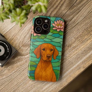May include: A phone case featuring a Vizsla dog illustration set against a stained-glass style background of lily pads and flowers in shades of green, pink, and blue. The case is on a wooden surface.