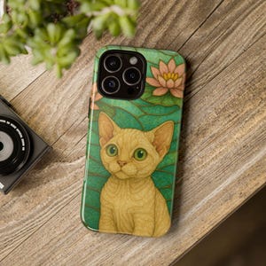 May include: A phone case featuring a detailed illustration of a golden cat with large green eyes. The case has a teal background with a lotus flower design. The phone case is on a wooden surface.
