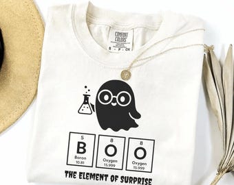 May include: Cream-colored t-shirt with a black ghost graphic wearing glasses and holding a beaker. Below the ghost are the letters B, O, and O, representing the word "BOO" in the periodic table. The text "THE ELEMENT OF SURPRISE" is below the letters.
