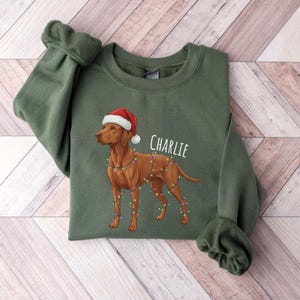 Op de afbeelding: Een olijfgroene sweatshirt met een bruine hond met een kerstmuts en lichtsnoer. De naam van de hond, "Charlie", is in het wit gedrukt. De sweatshirt is op een licht houtnerf oppervlak gevouwen.