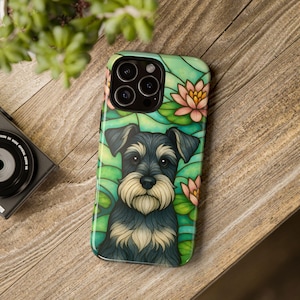 May include: A smartphone case featuring a Schnauzer dog design. The case has a colorful illustration of the dog surrounded by lily pads and pink lotus flowers. The case is on a wooden surface.
