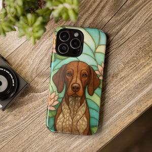May include: A phone case featuring a brown and white dog with long ears, set against a stained-glass style background of teal, green, and pink. The case is on a wooden surface, with a camera and plant in the background.
