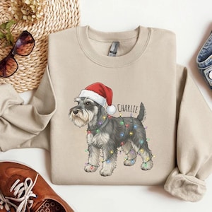 Puede incluir: Sudadera beige de cuello redondo con un perro Schnauzer con gorro de Papá Noel y luces navideñas. El nombre del perro, "Charlie", está impreso en negro. La sudadera es de color neutro, adecuada para la temporada navideña.