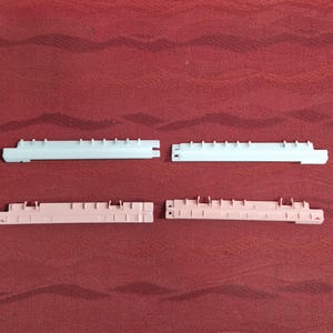 May include: Four plastic binder clips in the image. Two are white, and two are a light pink color. Each clip has a series of small, evenly spaced tabs along the top edge and a series of small rectangular tabs along the bottom edge.