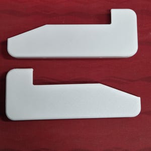 May include: Two white, L-shaped shelf brackets. Each bracket has a rectangular body with a right-angle extension. The brackets are made of a solid, opaque material and are set against a red background.