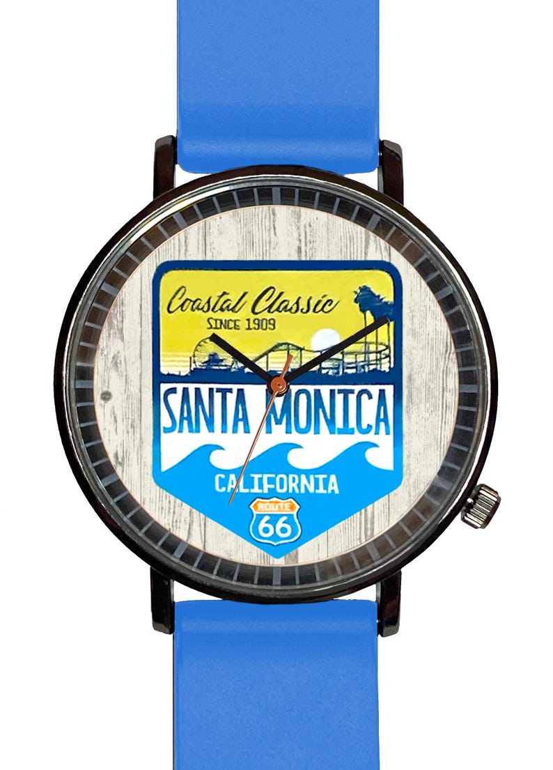 May include: A blue wristwatch with a black metal strap and a white face featuring a vintage-style graphic of the Santa Monica Pier with the text "Coastal Classic Since 1909 Santa Monica California Route 66".