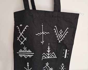 May include: Black tote bag with white geometric designs. The bag has two black straps and is hanging against a white wall. The designs are various shapes and symbols.