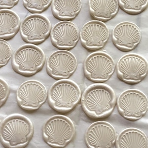 K&ouml;nnte beinhalten: Eine Ansammlung von cremewei&szlig;en Wachssiegeln, die jeweils ein detailliertes Muscheldesign aufweisen. Die Siegel sind eng angeordnet und bilden ein Muster. Das Muscheldesign umfasst strahlenf&ouml;rmige Linien und eine Schleife.
