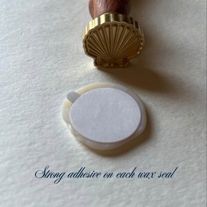 K&ouml;nnte beinhalten: Ein muschelf&ouml;rmiger Wachssiegelstempel mit Holzgriff, neben einem Wachssiegel mit wei&szlig;em Klebeaufkleber. Der Text "Strong adhesive on each wax seal" ist sichtbar.