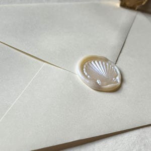 K&ouml;nnte beinhalten: Elfenbeinfarbener Umschlag mit einem perlmuttfarbenen Wachssiegel in Muschelform versiegelt. Das Siegel ist mit einem detaillierten Muscheldesign gepr&auml;gt. Ein Muschelwachsstempel ist im Hintergrund zu sehen.