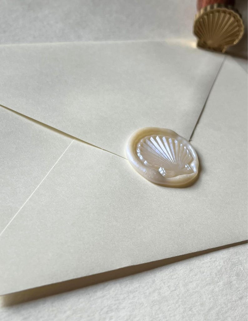 K&ouml;nnte beinhalten: Elfenbeinfarbener Umschlag mit einem perlmuttfarbenen Wachssiegel in Muschelform versiegelt. Das Siegel ist mit einem detaillierten Muscheldesign gepr&auml;gt. Ein Muschelwachsstempel ist im Hintergrund zu sehen.