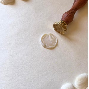 K&ouml;nnte beinhalten: Ein Wachssiegel mit Muschelmuster, mit einem Holzgriff und einem goldfarbenen Siegelkopf. Ein Muschelabdruck ist sichtbar. Muscheln sind auf einer cremefarbenen Oberfl&auml;che verstreut.