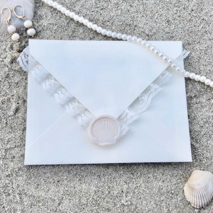 K&ouml;nnte beinhalten: Ein wei&szlig;er Umschlag mit Spitzenbesatz und einem Muschel-Wachssiegel, auf Sand. Eine Perlenkette und goldene Ohrringe mit wei&szlig;en Perlen sind ebenfalls vorhanden, was auf ein Strand- oder Hochzeitsthema hindeutet.