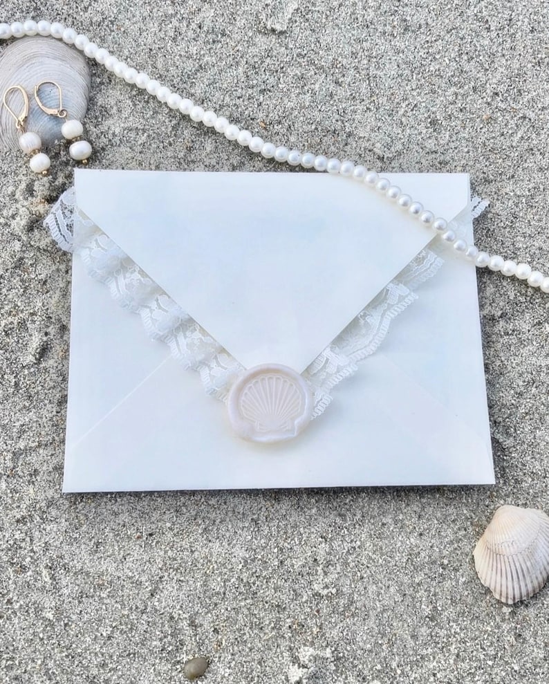 K&ouml;nnte beinhalten: Ein wei&szlig;er Umschlag mit Spitzenbesatz und einem Muschel-Wachssiegel, auf Sand. Eine Perlenkette und goldene Ohrringe mit wei&szlig;en Perlen sind ebenfalls vorhanden, was auf ein Strand- oder Hochzeitsthema hindeutet.