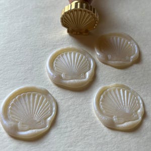 K&ouml;nnte beinhalten: Ein Muschel-Wachssiegelstempel mit Holzgriff und vier Muschel-Wachssiegeln. Der Stempel hat einen goldfarbenen Metallkopf. Die Wachssiegel sind cremefarben mit einem perlmuttartigen Schimmer, der Hintergrund ist hellbeige.
