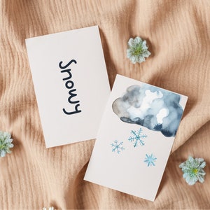Op de afbeelding: Twee witte kaarten met aquarel illustraties. Eén kaart leest "Snowy" in zwart schrift. De andere heeft een blauwe en grijze aquarel wolk met drie blauwe sneeuwvlokken. De kaarten liggen op een beige stof.