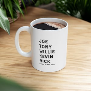 May include: White ceramic mug filled with dark coffee. The mug features the names JOE, TONY, WILLIE, KEVIN, and RICK in black text, with "LIVING IN KEY WEST" below. The mug sits on a wooden surface.